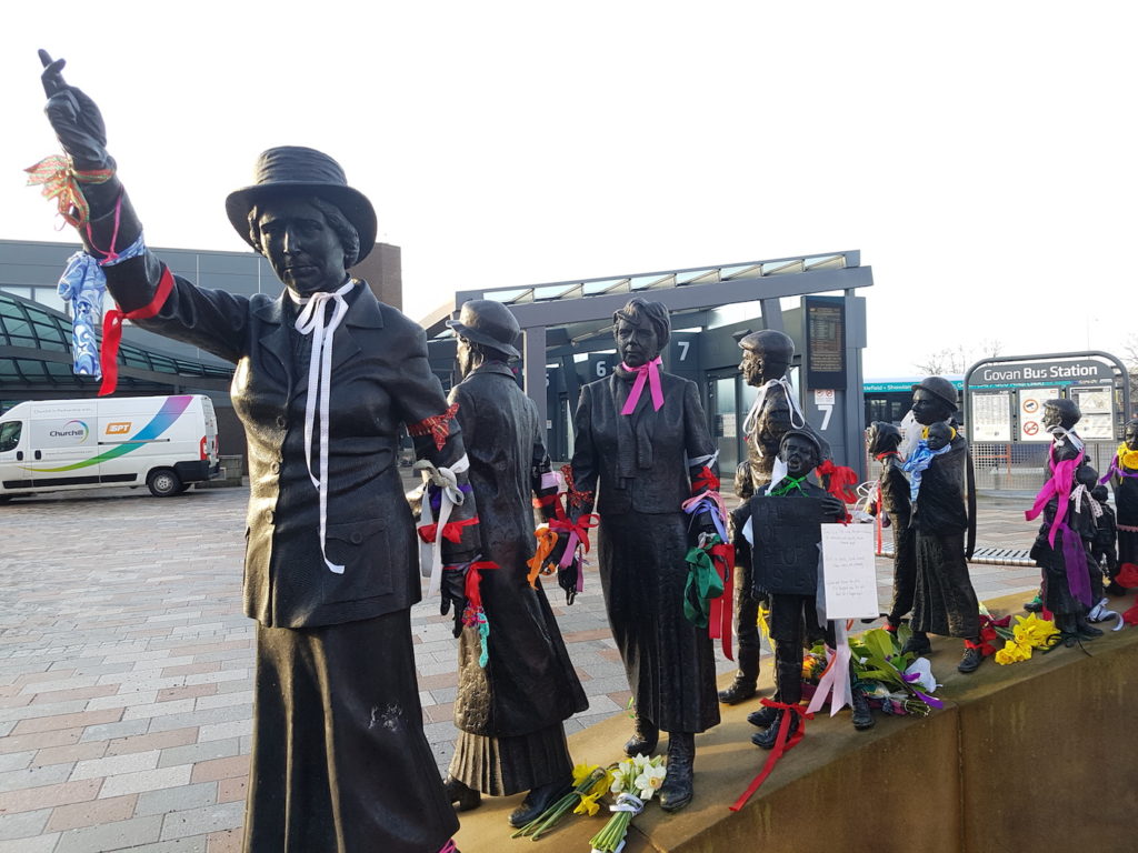 A photograph of a sculpture of Mary Barbour, a female figure holding one arm in the air, with a group of men women and children behind her, decorated with ribbons and flowers