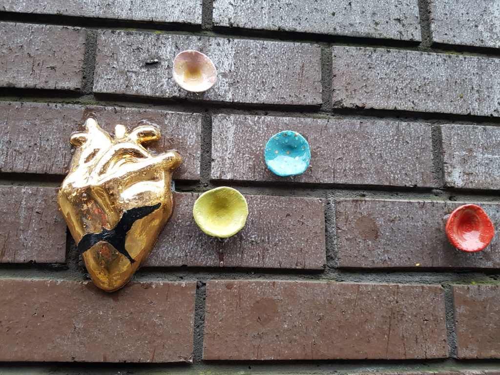 A photograph of a brick wall with colourful ceramic objects stuck to it; a golden heart with a black crack across it, yellow, blue, pink and red circles.