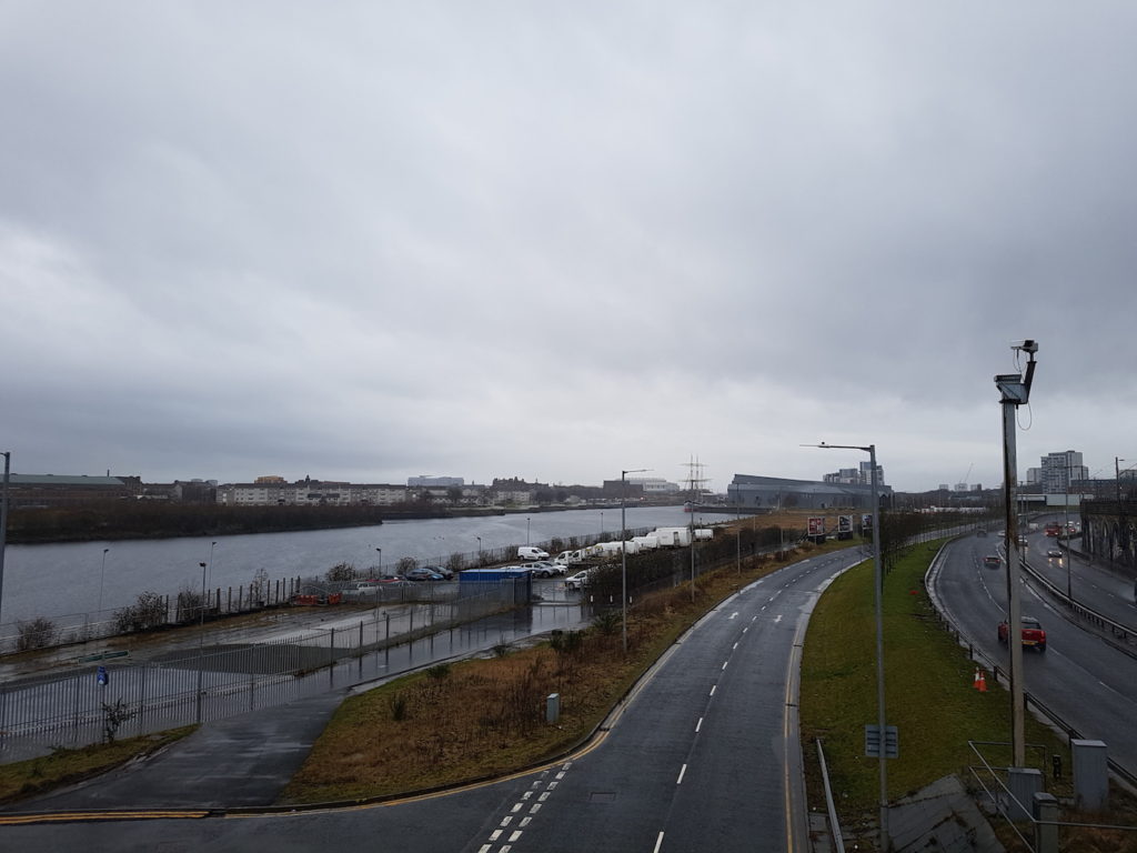 A photograph of a grey river with a car park alongside and then 6 lanes of road with cars driving on them. A CCTV camera on a tall pole in the right of the frame.