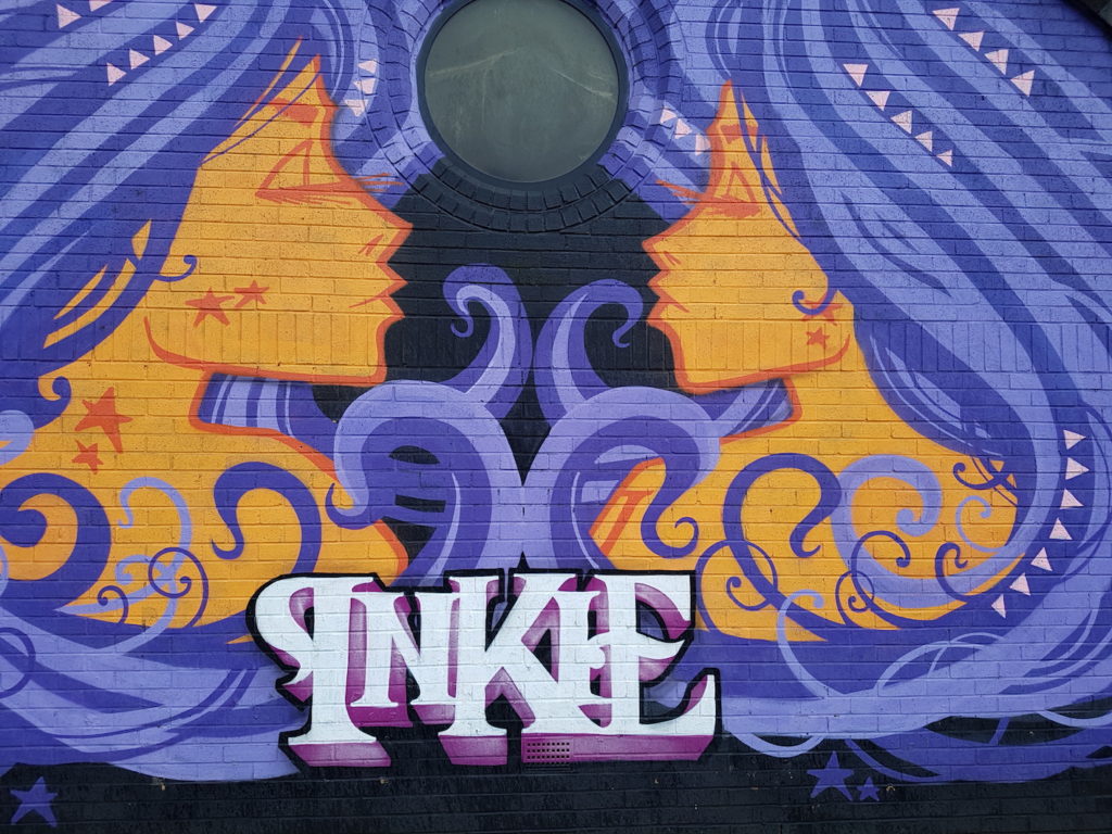 A photograph of a wall mural with two women facing each other. They are painted with yellow and ornage skin and purple/blue hair curling around them in stylised swirls. The word 'Inkie' is painted in large white letters at the bottom.