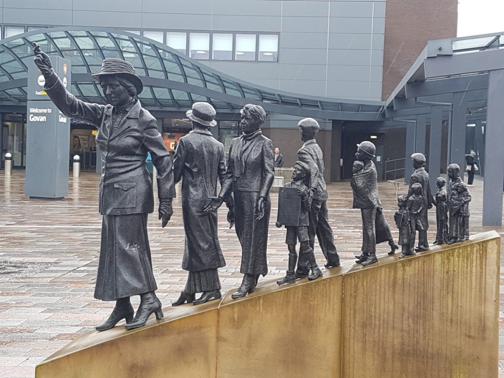 A photograph of a sculpture of figures, the Govan rent strikers, with the figure of Mary Barbour holding her hand in the air at the front.