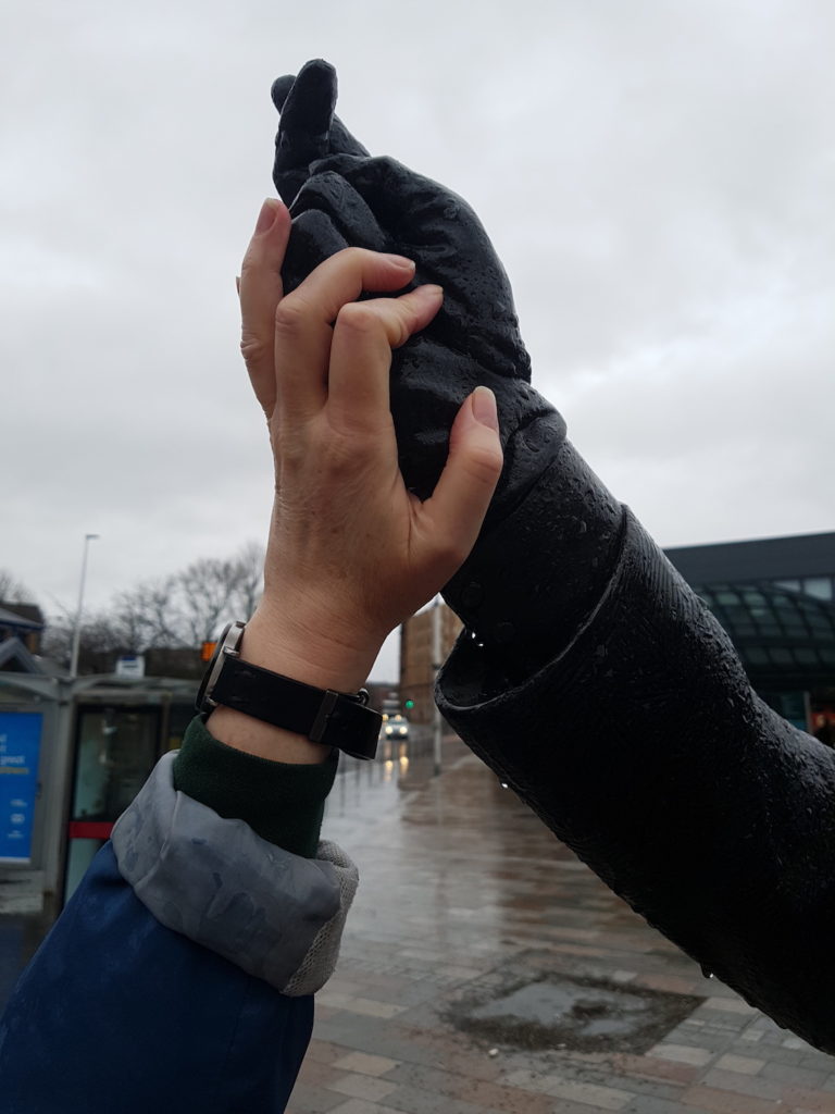 A photograph of the authors hand held up to meet the hand of the sculpture of Mary Barbour